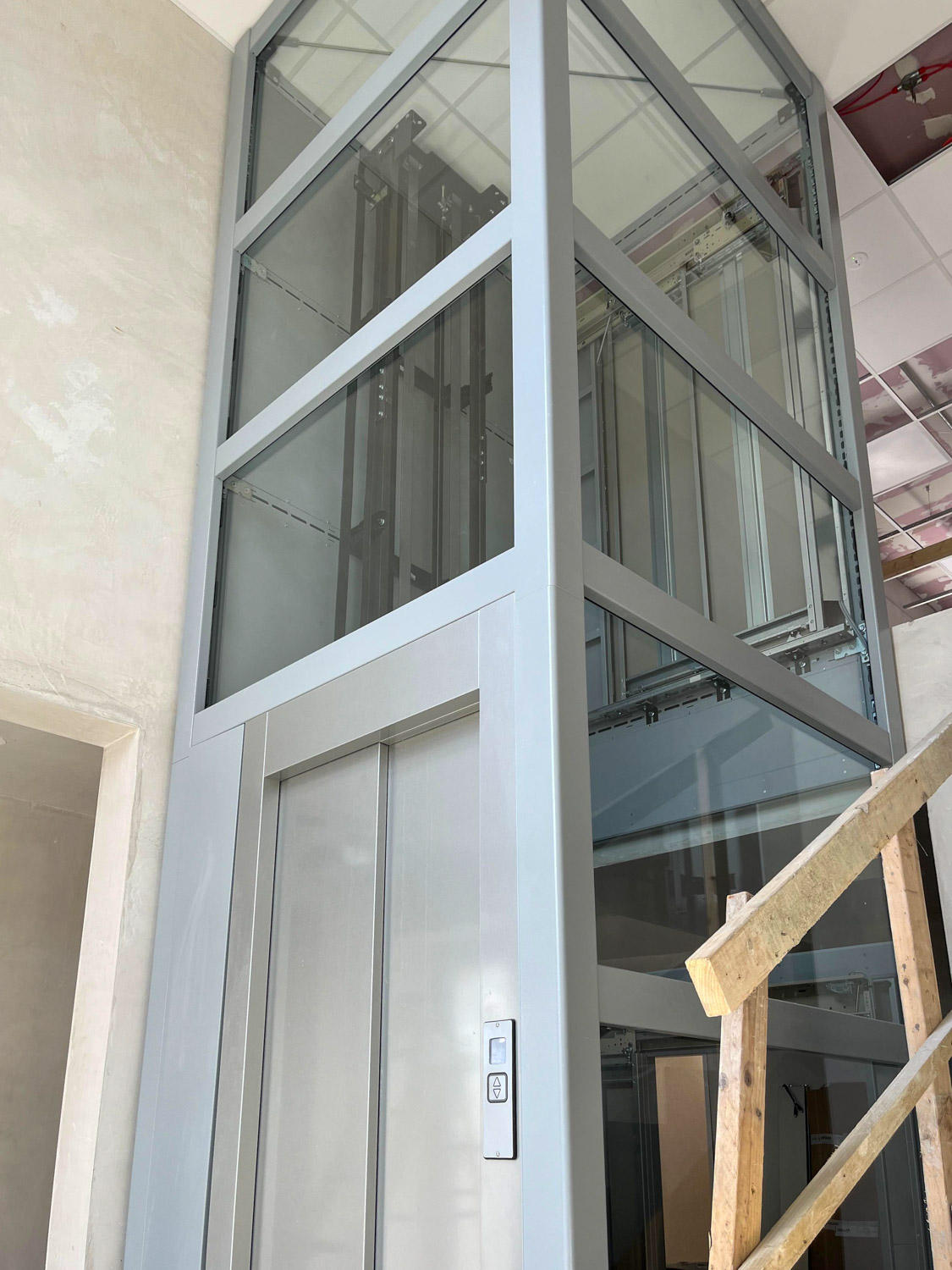 Modern glass and metal public elevator shaft with closed silver elevator doors and control panel in a building interior under construction, with wooden scaffolding nearby.