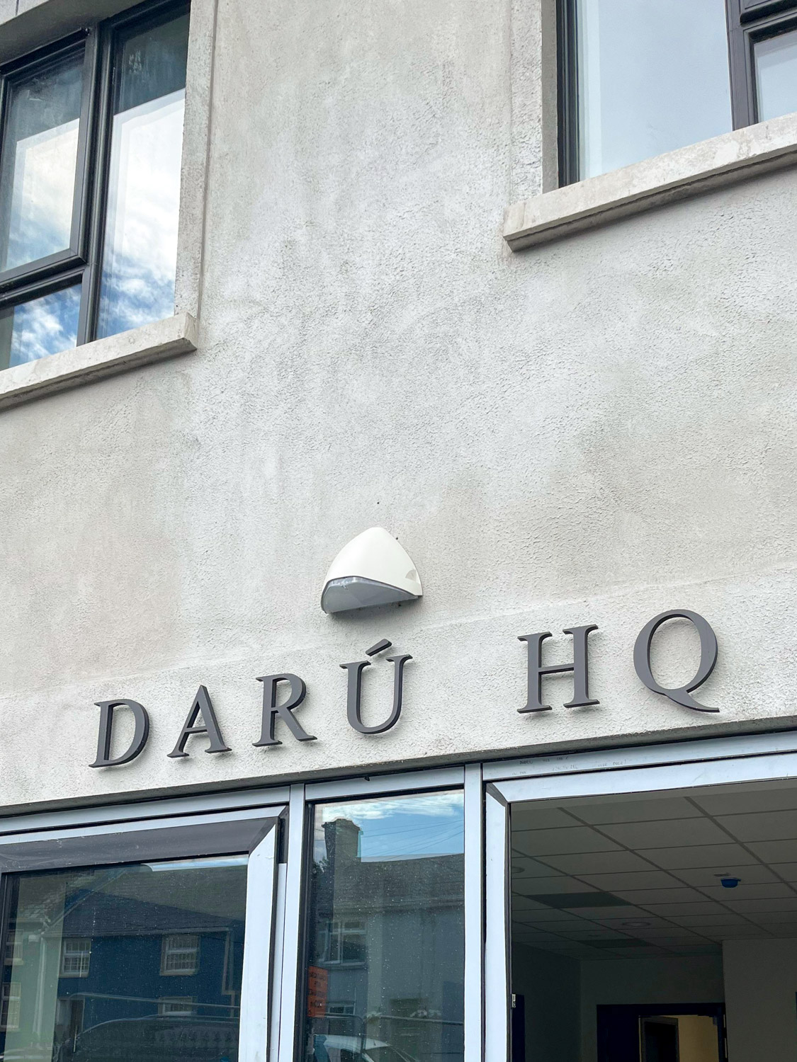 Exterior of a building with gray textured walls, two windows at the top, and a glass door below with reflected houses. Above the door is a black sign reading 'DARÚ HQ' with a small white light fixture mounted above the sign.