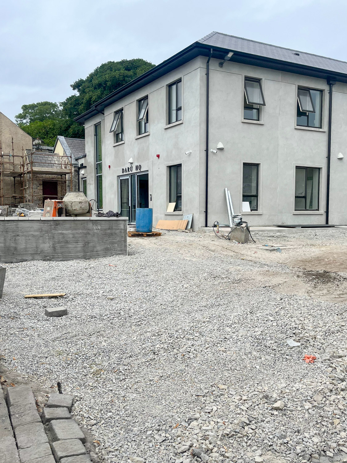 Two-story grey building with several windows and a door labeled 'DARÚ HQ' under construction, with gravel and construction materials in the foreground.