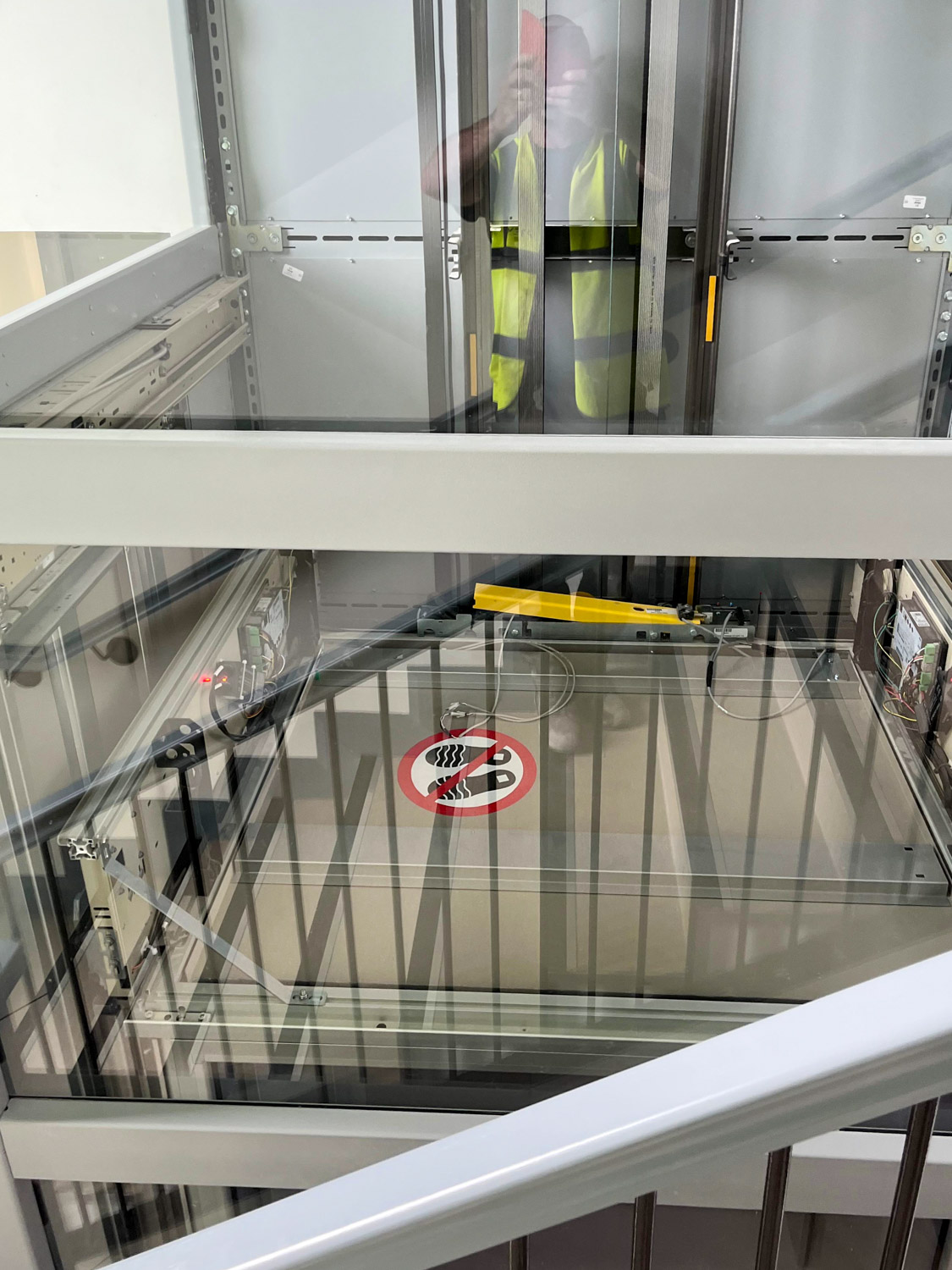 View of an elevator shaft with metal framework and a no smoking sign on a glass platform, reflecting a person in a yellow safety vest taking the photo.