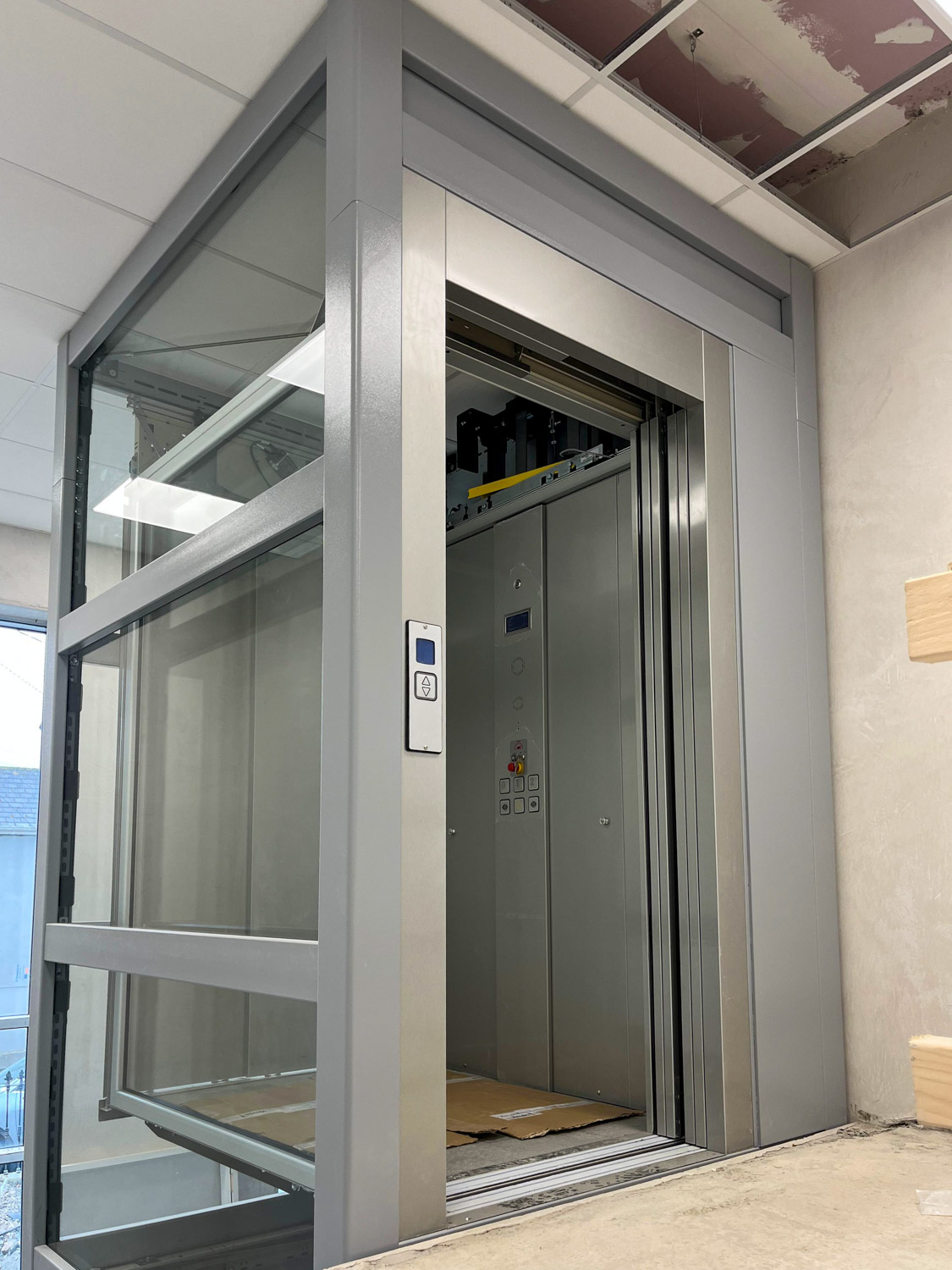 Modern indoor elevator with glass walls and metal frame, open door showing the cabin with control panel and cardboard on the floor.