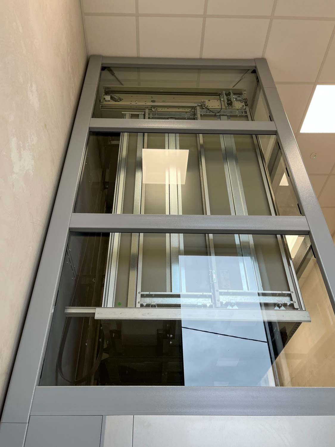 Interior view of a public elevator shaft showing metal framework and mechanical components behind a glass panel inside a building with beige walls and a tiled ceiling.