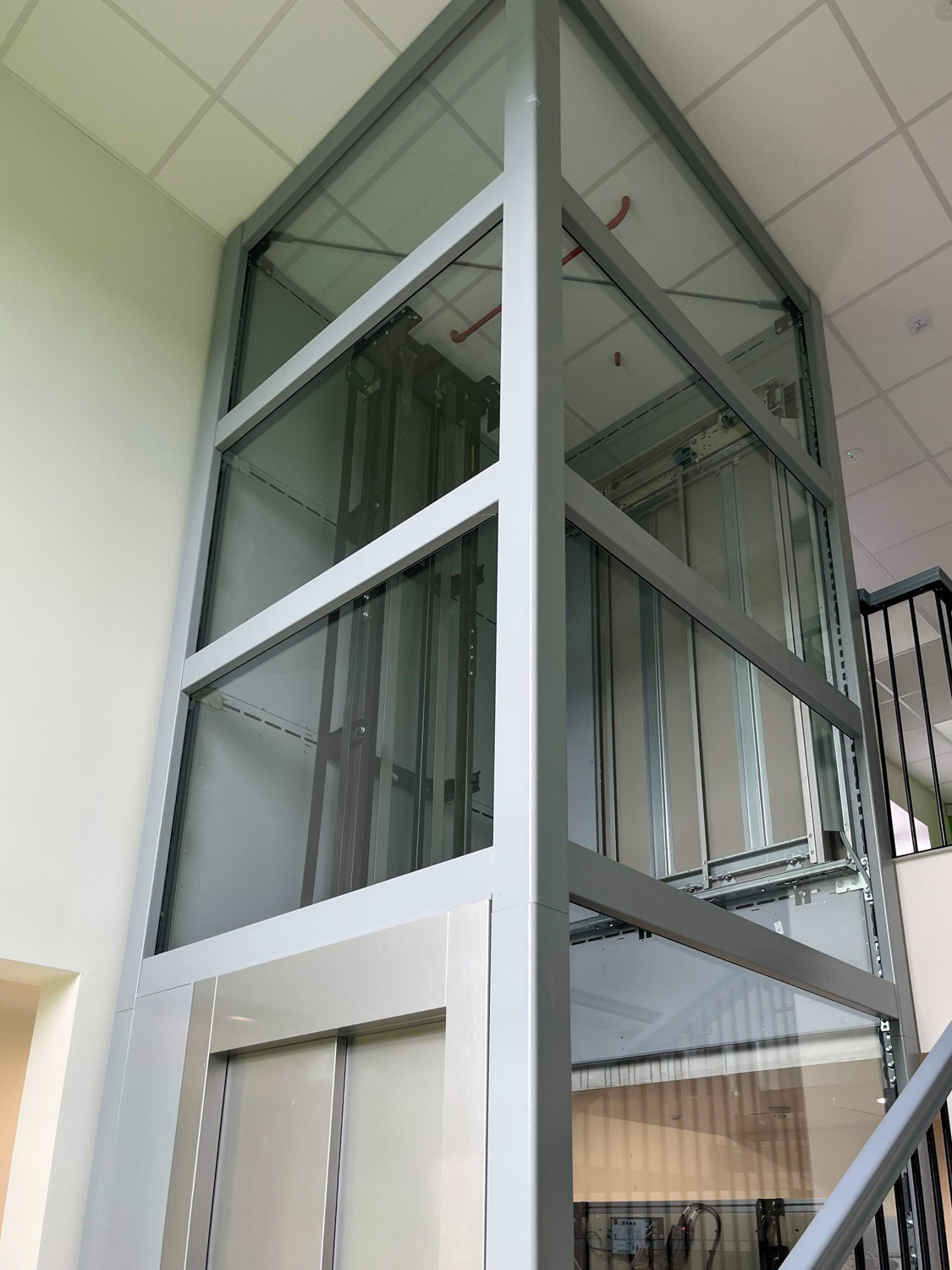 Modern glass and metal elevator shaft inside a building, showing the elevator machinery and doors at the bottom floor.