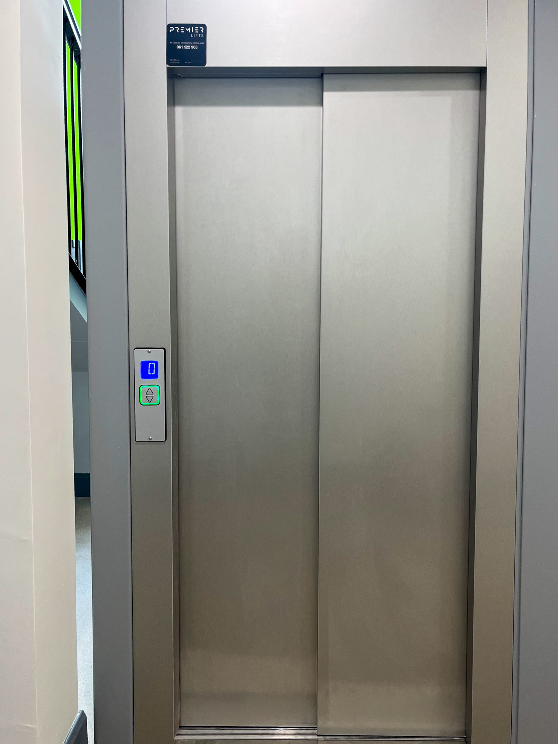 Closed stainless steel elevator doors with a lit blue floor indicator showing 0 and a green-bordered call button panel on the left side.
