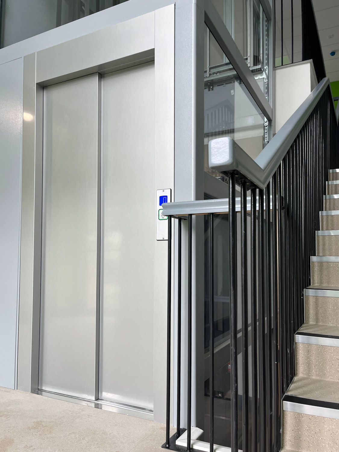 Eco public elevator with closed silver doors next to a staircase with black metal railing and beige steps.