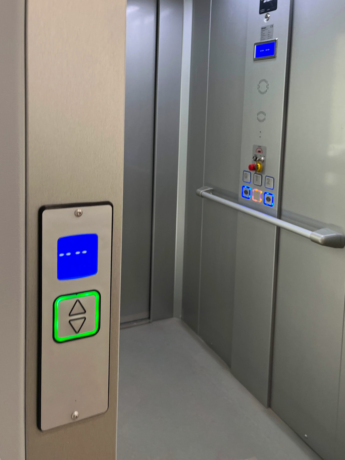 Interior of a grey metal eco public lift with control panel showing illuminated floor buttons and a horizontal handrail on the right wall, and the outside elevator call button panel on the left with up and down arrows surrounded by green light.
