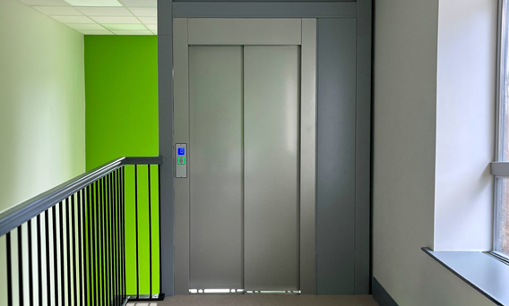 Closed grey eco public lift doors in a hallway with green and white walls and a black railing on the left side next to a window on the right.