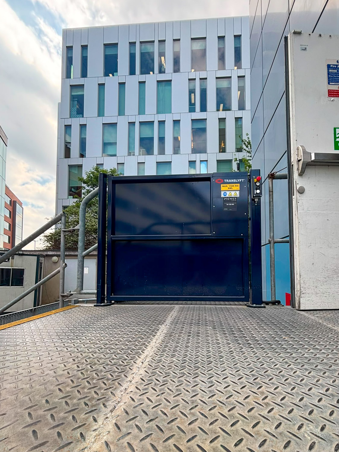 View from a metal platform with diamond plate pattern facing a closed dark blue scissor lift safety gate labeled Translyft with a control panel on the right, outside a modern multi-story building with vertical white and glass panels.