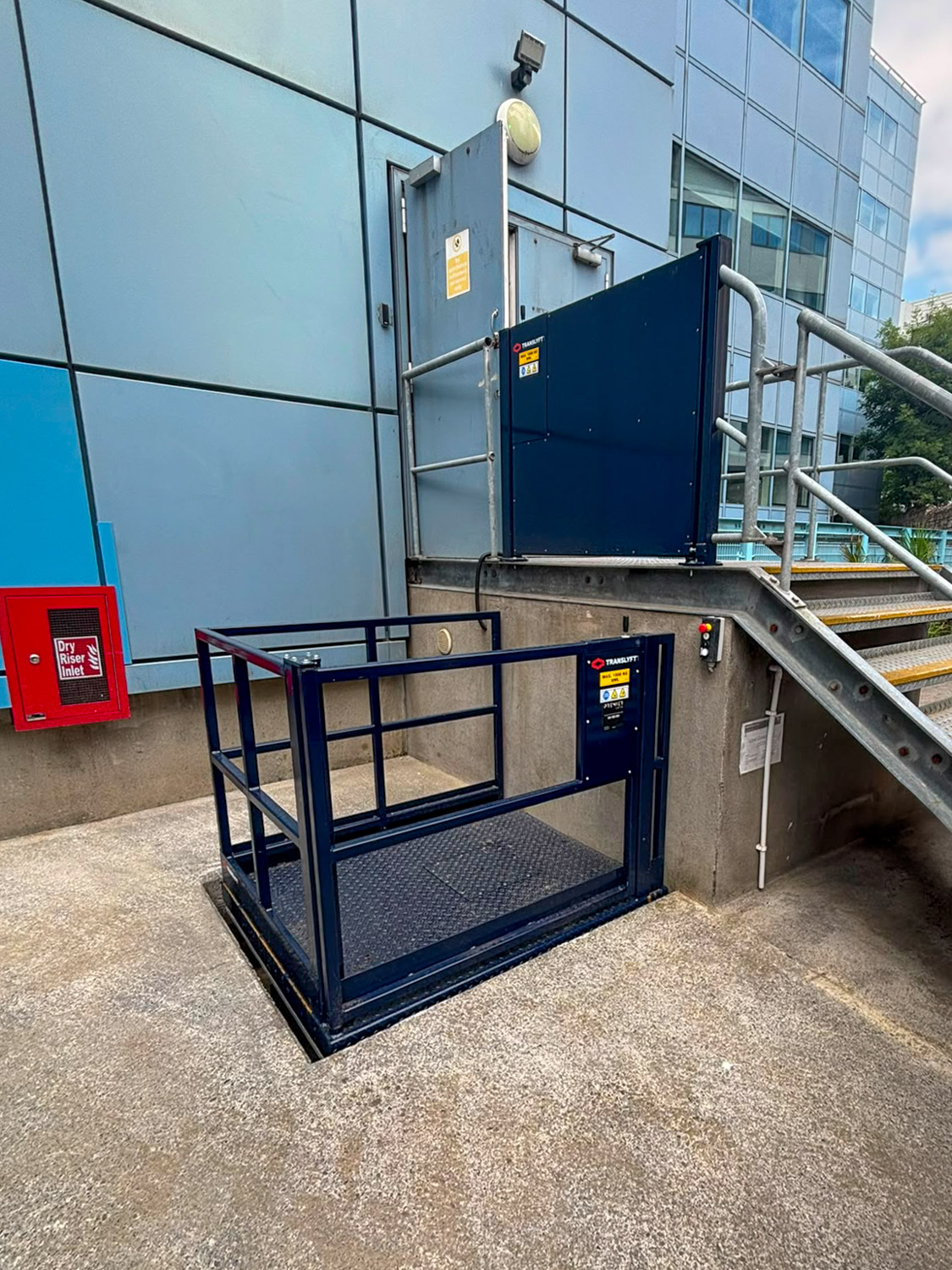 Outdoor scissor lift in front of stairs beside a modern glass and metal building exterior, with a closed blue door and a red dry riser inlet box on the wall.