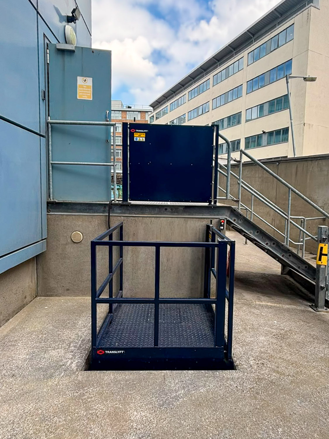 Outdoor scissor goods lift with navy blue metal safety rails and a closed gate, positioned at the edge of a concrete surface near stair railings and a blue building wall.