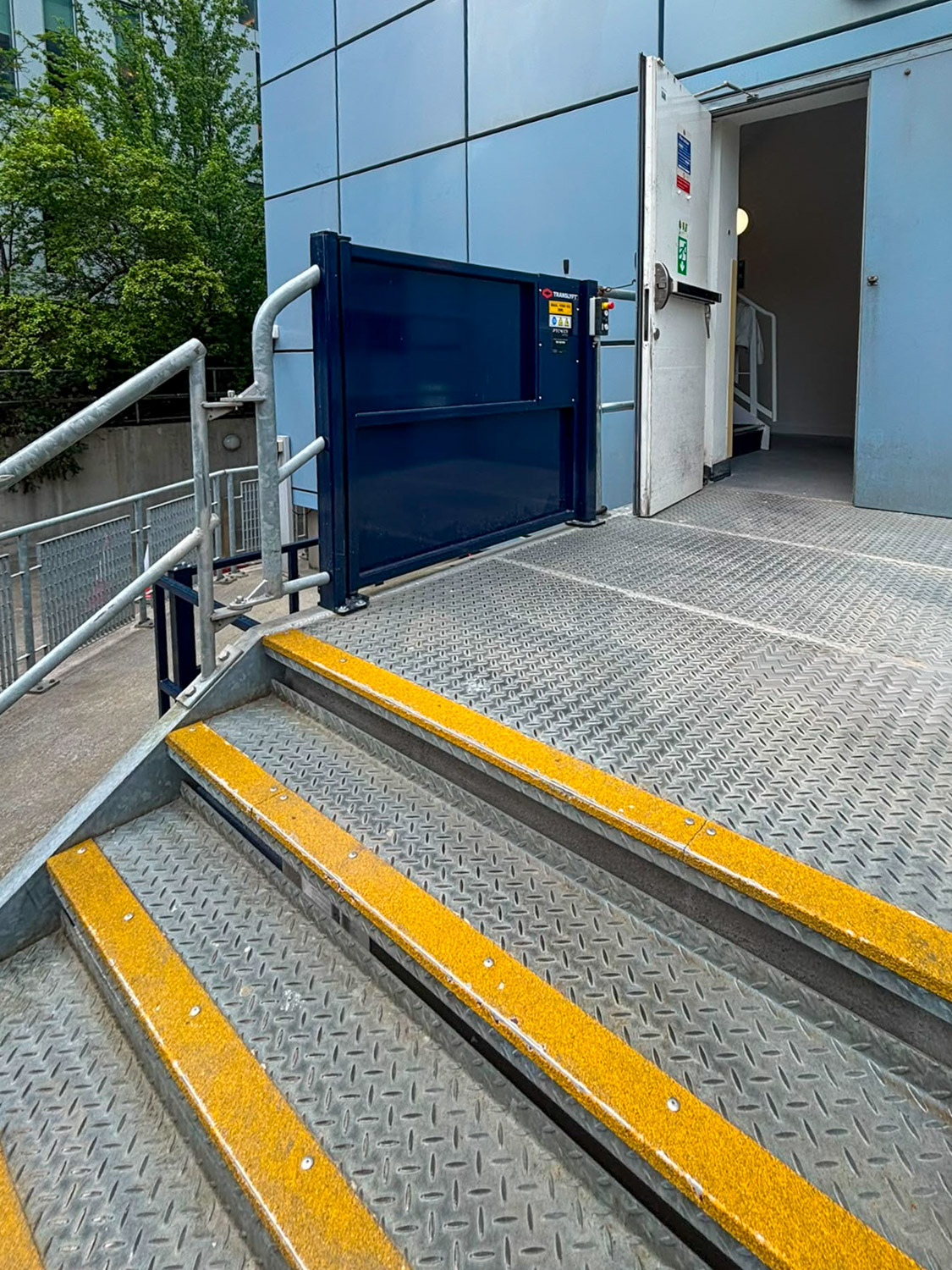Metal outdoor steps with yellow safety strips leading up to a metal platform with a blue goods scissor lift next to an open door of a modern building.