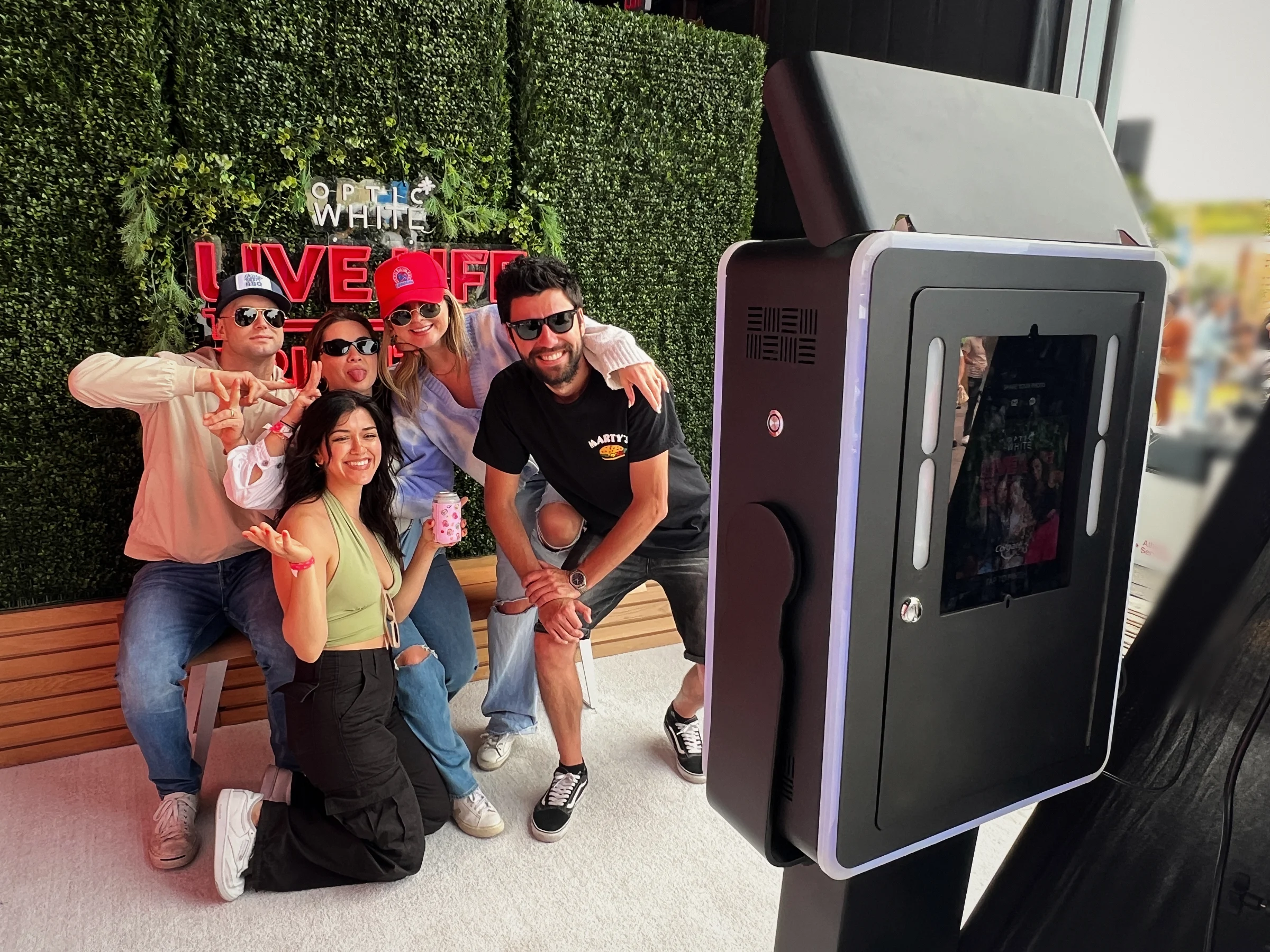 Guests posing at a Post Pop photo booth kiosk in front of a branded hedge wall at a Colgate Optic White activation