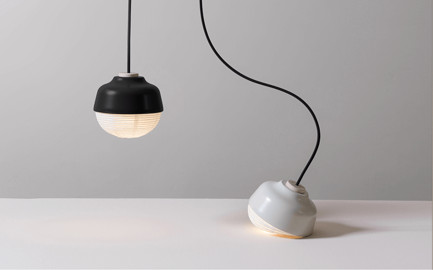 Two modern hanging pendant lights; one black suspended, one gray resting on a white surface.
