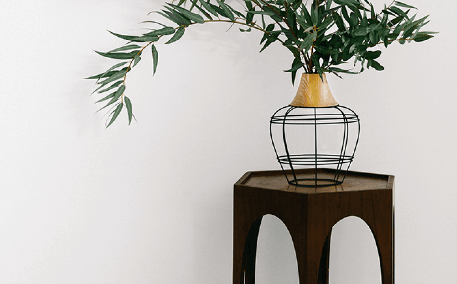 Modern wireframe vase with wooden neck holding green leafy branches on a small dark wood table.