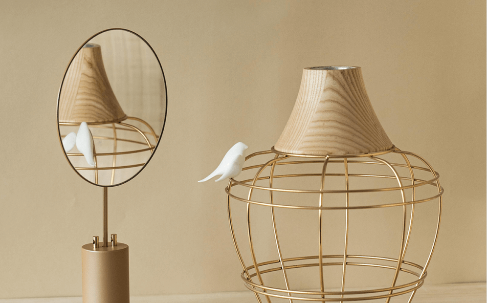Decorative gold wire frame with wooden top and white bird figurine beside oval mirror reflecting them.