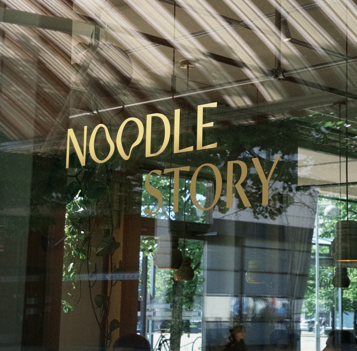 Glass window showing the words 'NOODLE STORY' with reflections of outdoor trees and street scene.