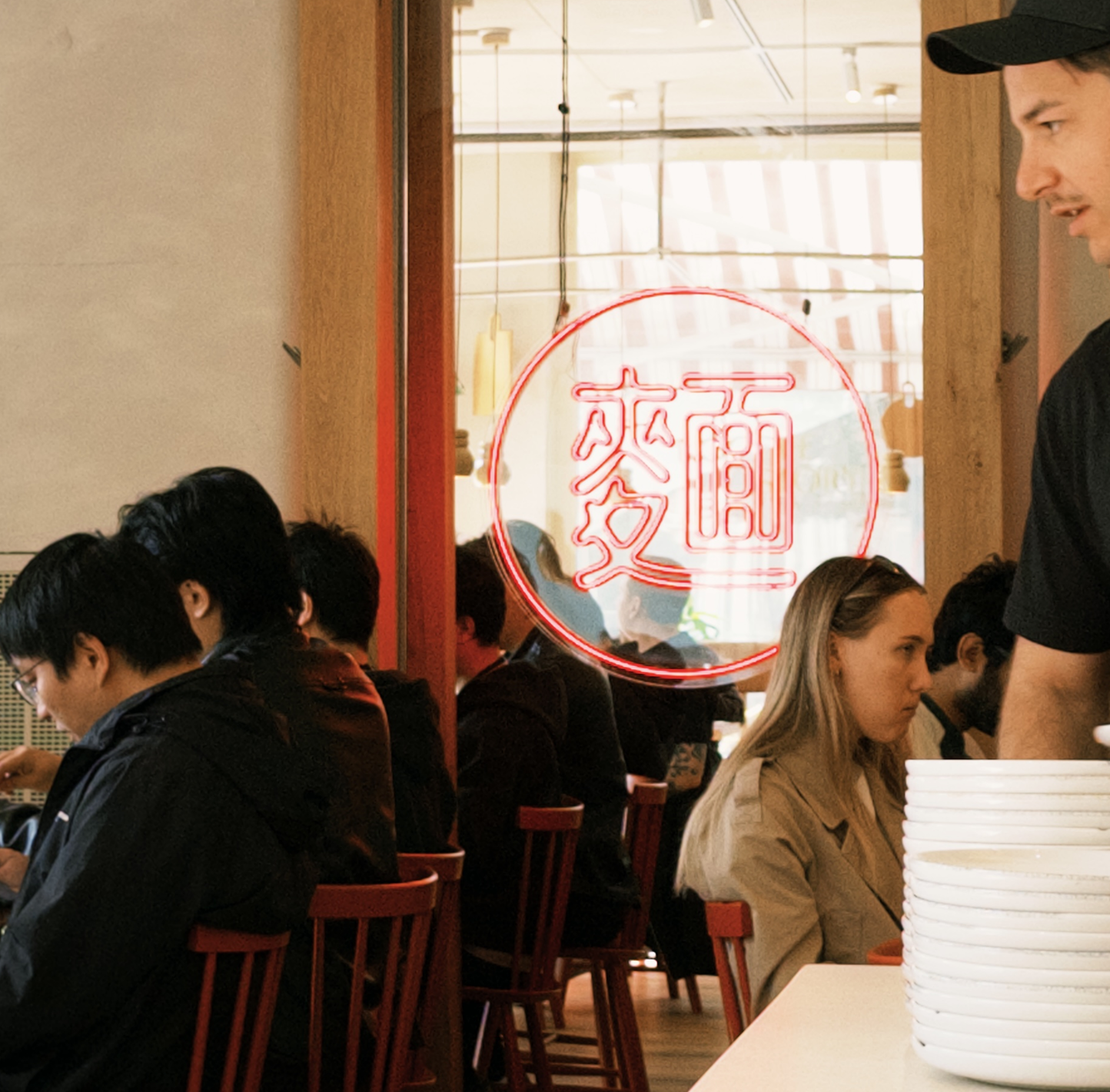 People seated in a restaurant with a neon sign and a stack of white bowls on the counter.