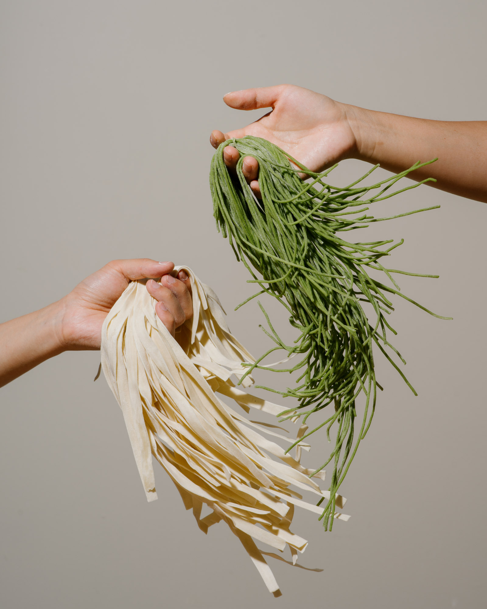 Two hands holding fresh uncooked pasta: one hand holds green pasta, the other holds white pasta.