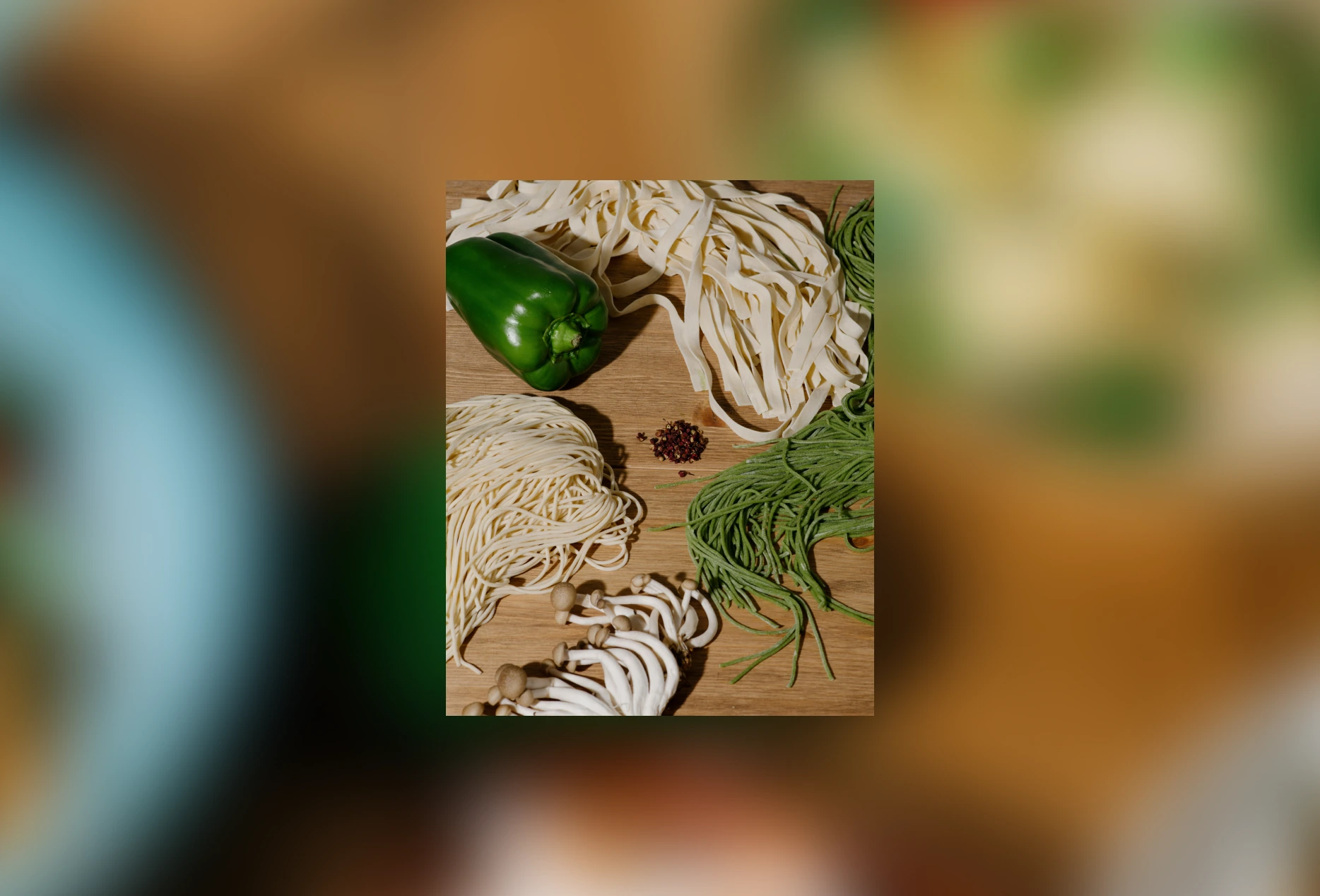 Green bell pepper, bundles of white and green noodles, mushrooms, and peppercorns on a wooden surface.