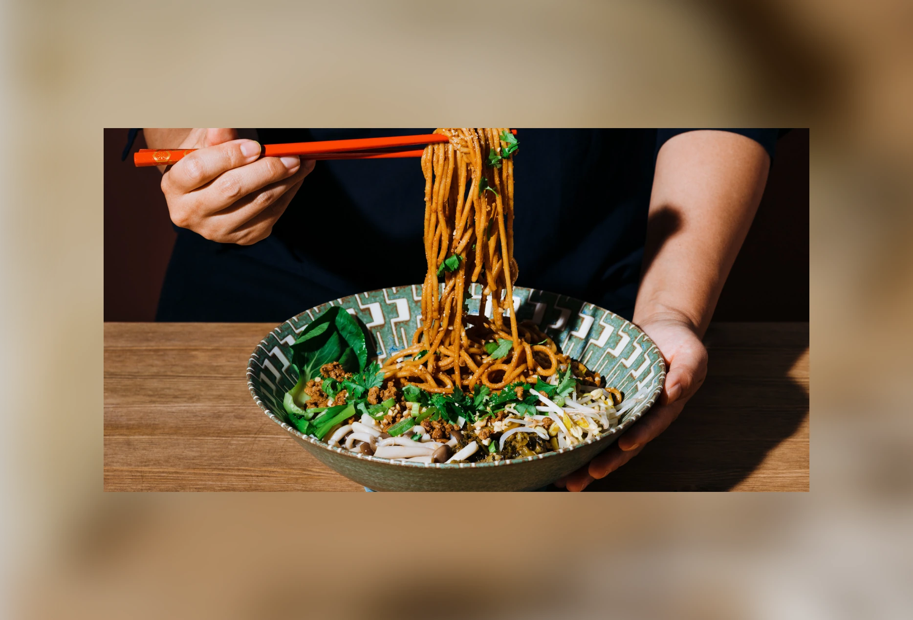 Hands holding bowl and red chopsticks lifting brown noodles with green vegetables and sprouts.