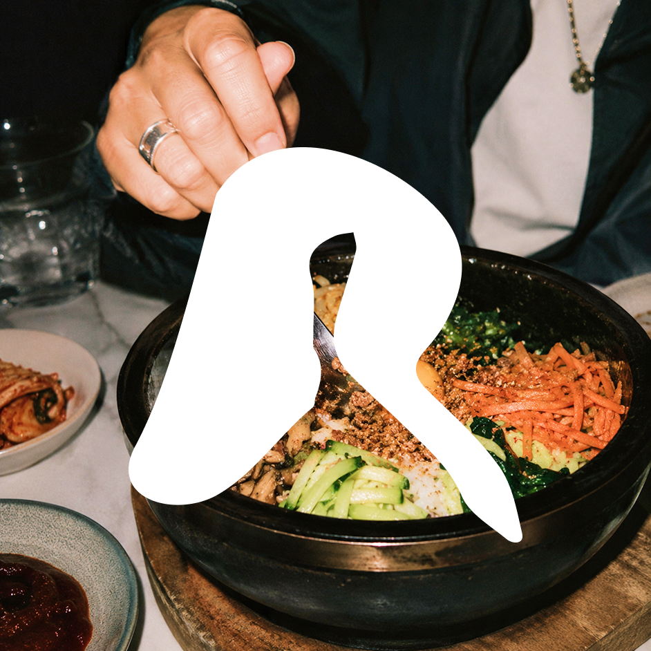 Hand holding chopsticks over a bowl of bibimbap with vegetables and rice on a table.