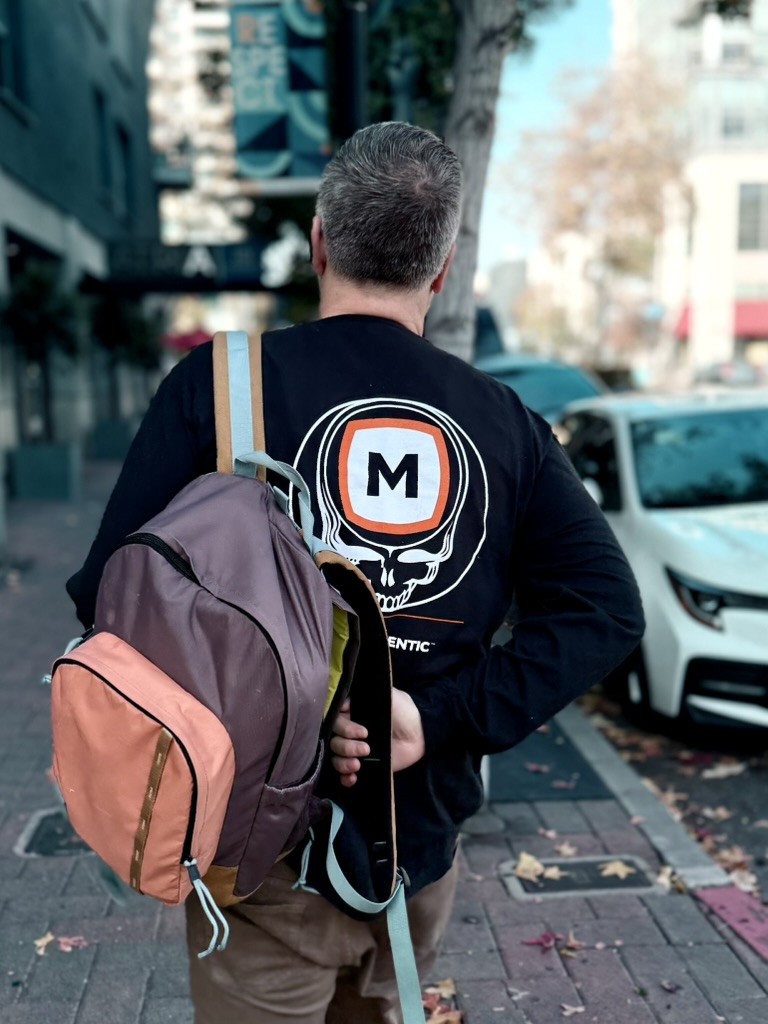 Man with gray hair walking on a city sidewalk carrying a purple and orange backpack over one shoulder and wearing a black shirt with a large 'M' skull logo on the back.