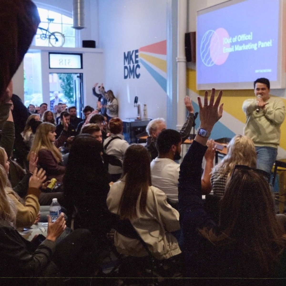 Audience members raising hands during a presentation titled 'Out of Office Email Marketing Panel' in a brightly lit room with MKE DMC logo on the wall.