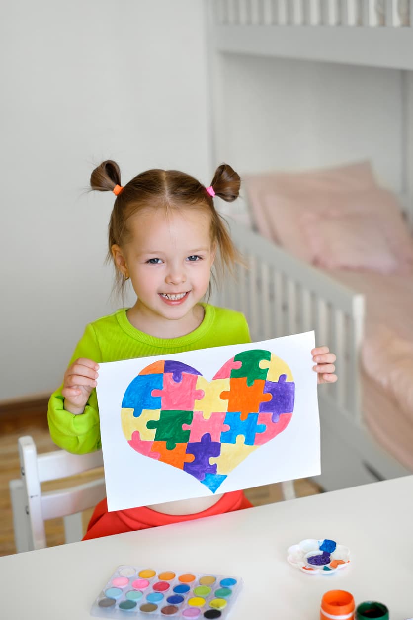 A smiling young child holds a colourful heart made of puzzle pieces during an arts and crafts activity. The image represents autism awareness, creative expression, neurodiversity, and child development in a positive, supportive environment.