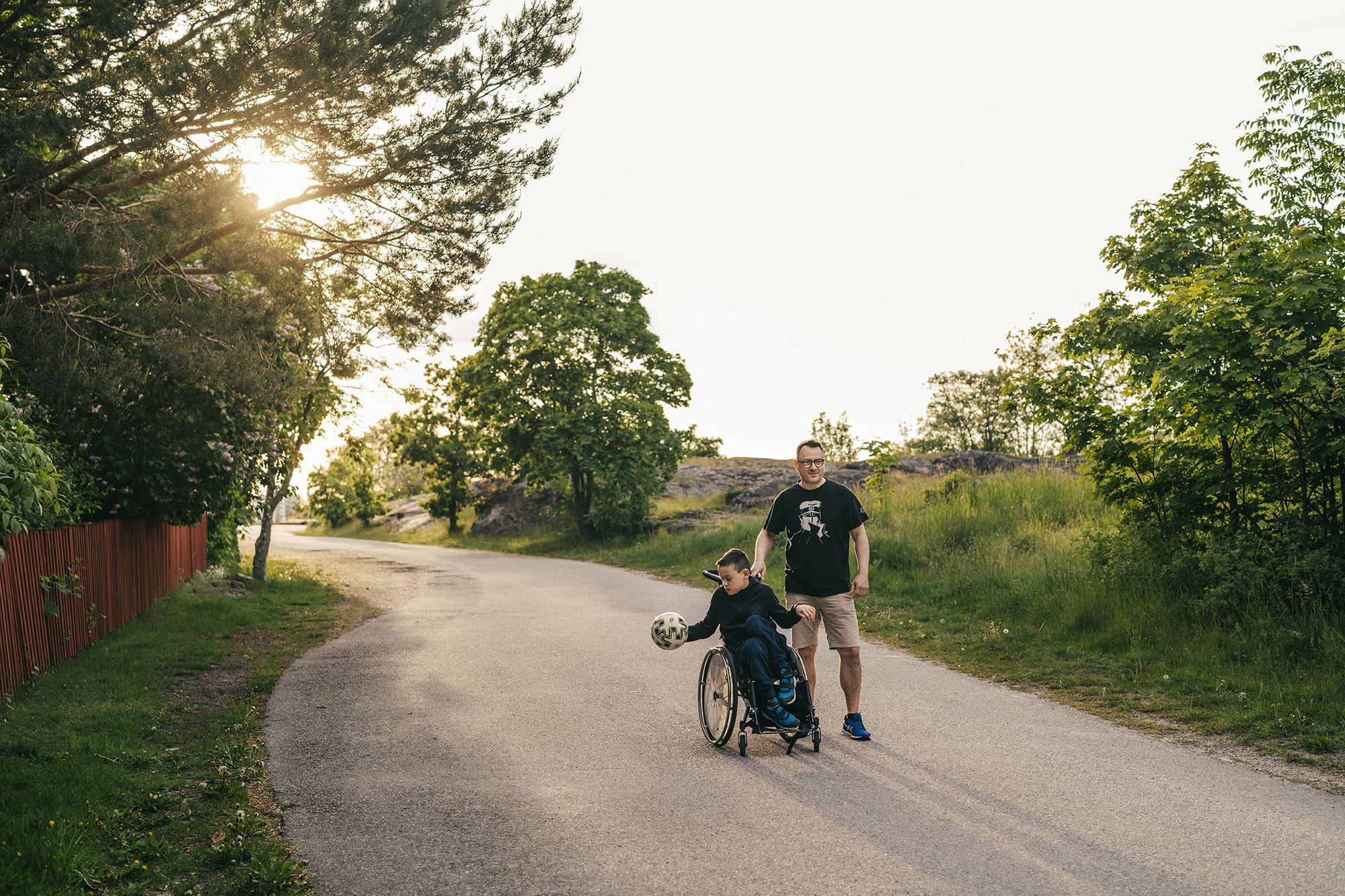 En pojke i rullstol tillsammans med en man på en parkväg. Det är sommar.
