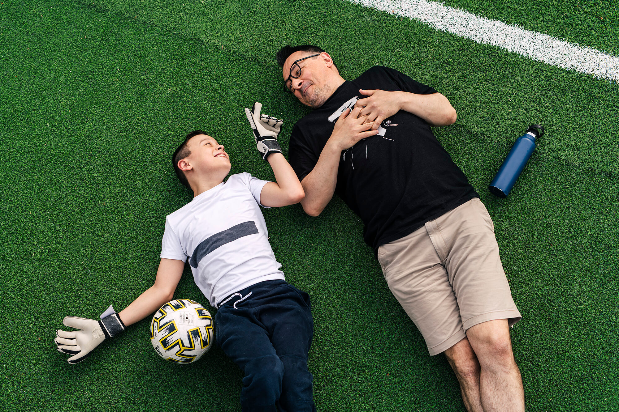 En pojke och en man ligger på rygg på en fotbollsplan.