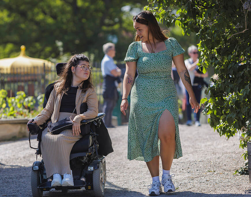 En rullstolsburen kvinna i beige kläder  bredvid en gående kvinna i grön klänning. De är i en solig park.