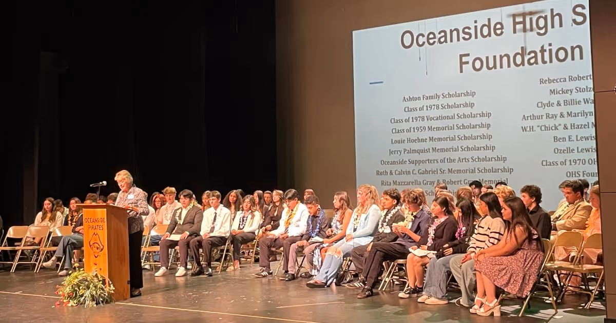 A speaker stands at a podium with an Oceanside Pirates logo addressing seated students on stage during an awards ceremony with scholarship names projected on a screen.