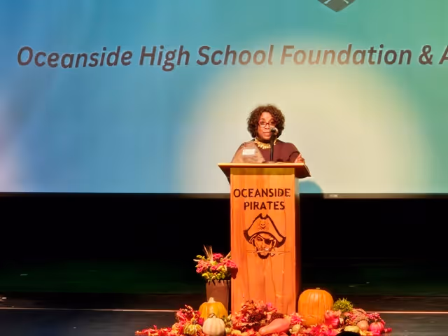 Woman speaking at a wooden podium with 'Oceanside Pirates' and pirate logo, with floral and pumpkin decorations at the base, in front of a screen displaying 'Oceanside High School Foundation'.
