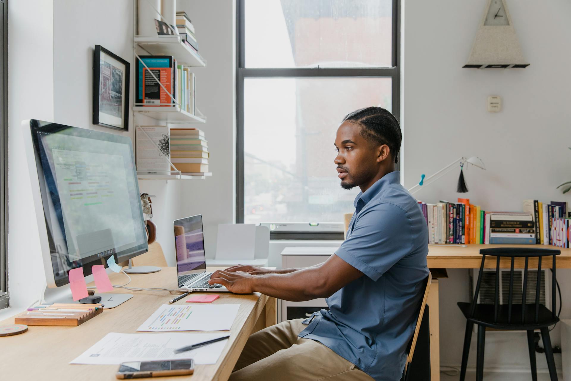 A writer sits at a desk, typing on a laptop and looking at a larger monitor.