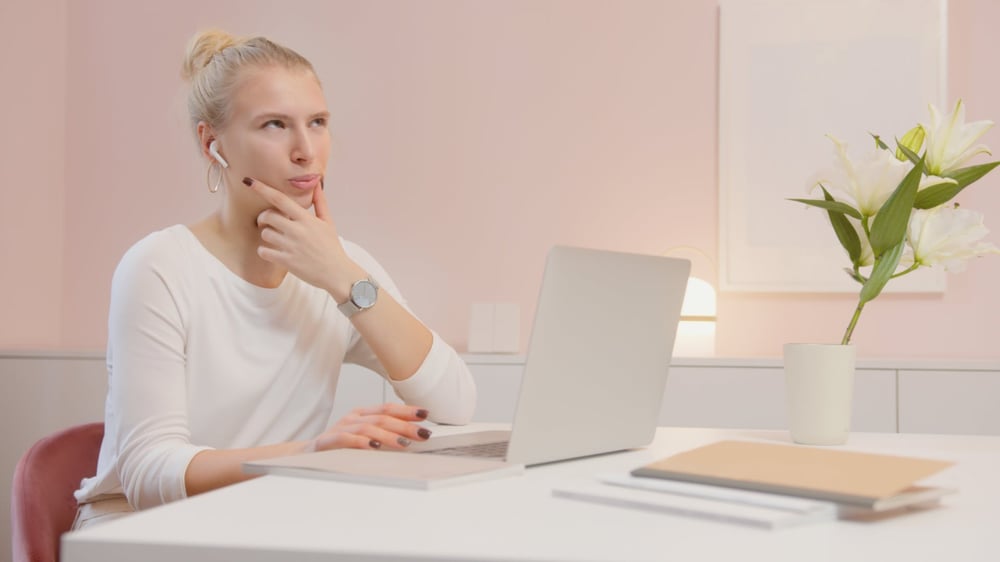 A writer in a white shirt sits at a white desk in front of a laptop, touching their face and thinking.
