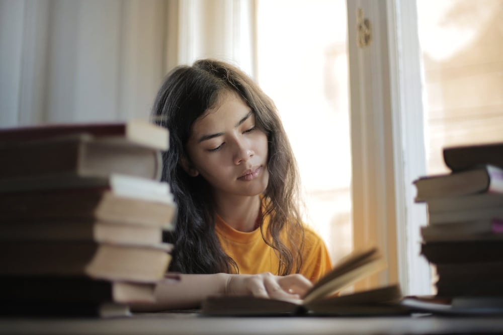 A person reads a book while sitting surrounded by stacks of other books.