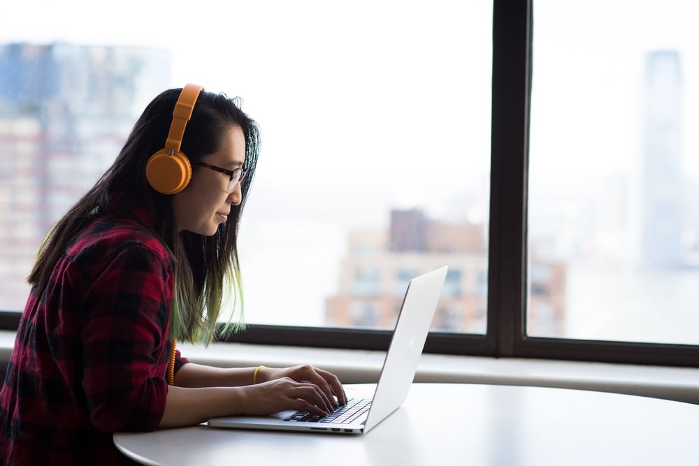 A person wearing headphones and typing on a laptop beside a window with a city view.