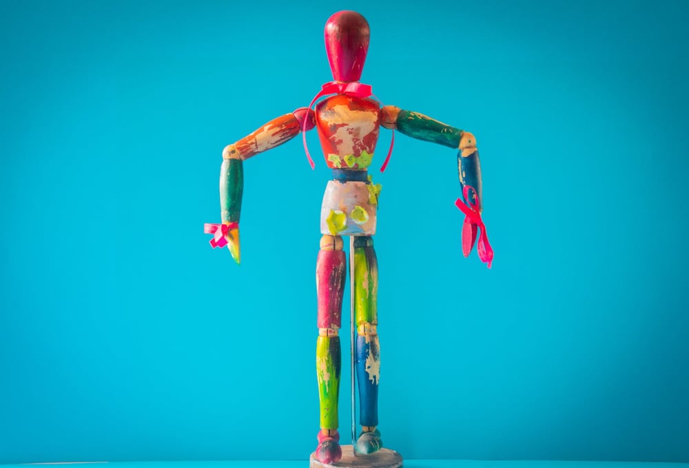 A wooden artist's mannequin that's been painted in several blotchy colors and decorated with ribbons.