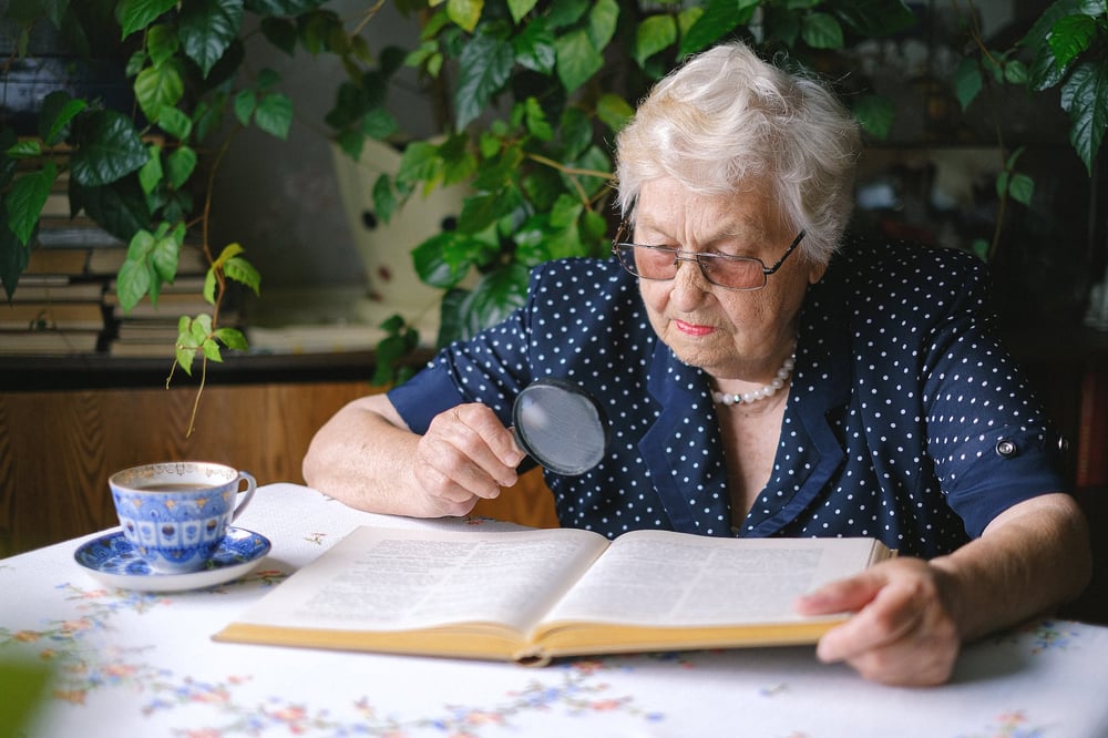 An elderly person sits at an outdoor table with a teacup, looking at a big book through a magnifying glass.