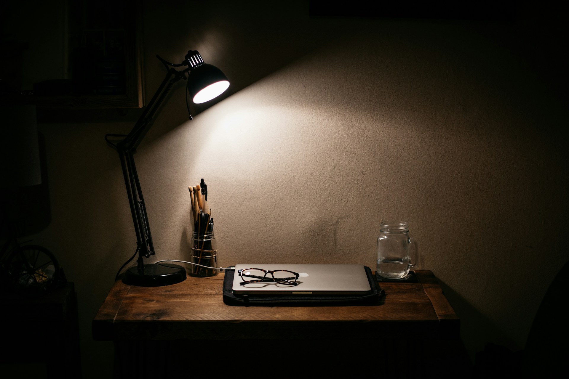 In a dark room, a small desk lamp illuminates a closed laptop, glasses, and a glass of water.