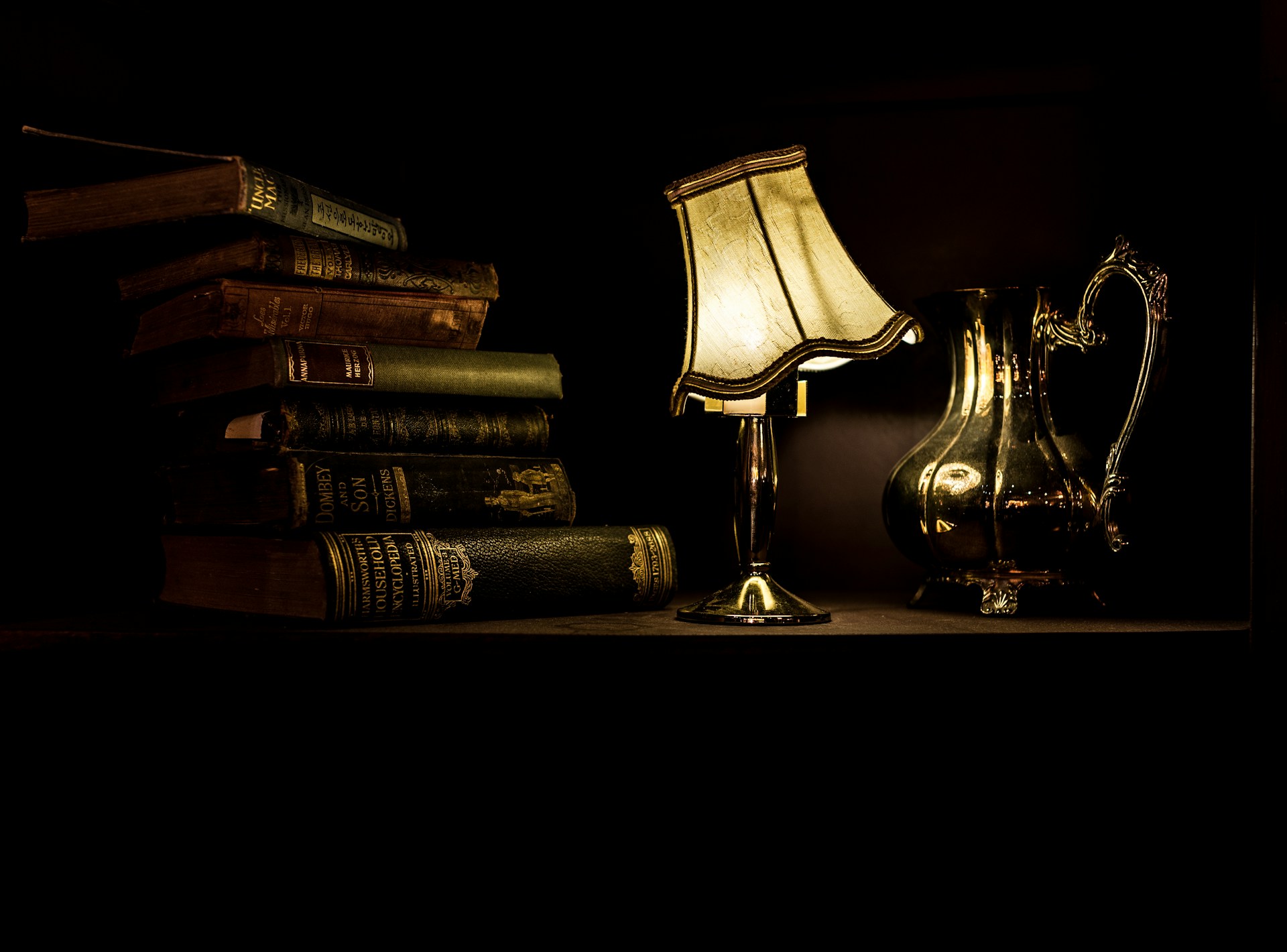 A stack of black and brown antique novels beside an antique lamp with its shade at a tilt.