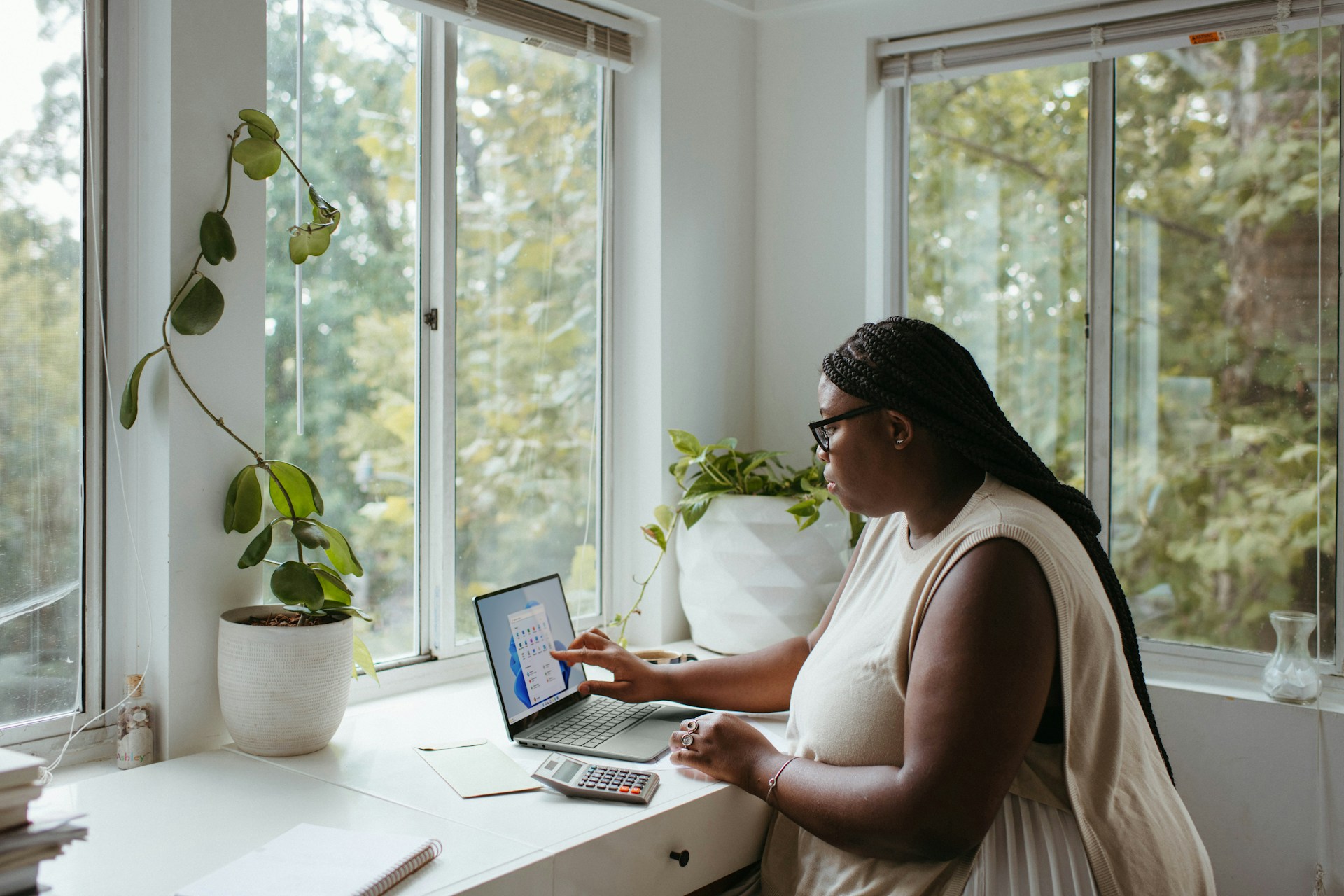 A person works on a computer at a desk beside large windows looking out on trees.