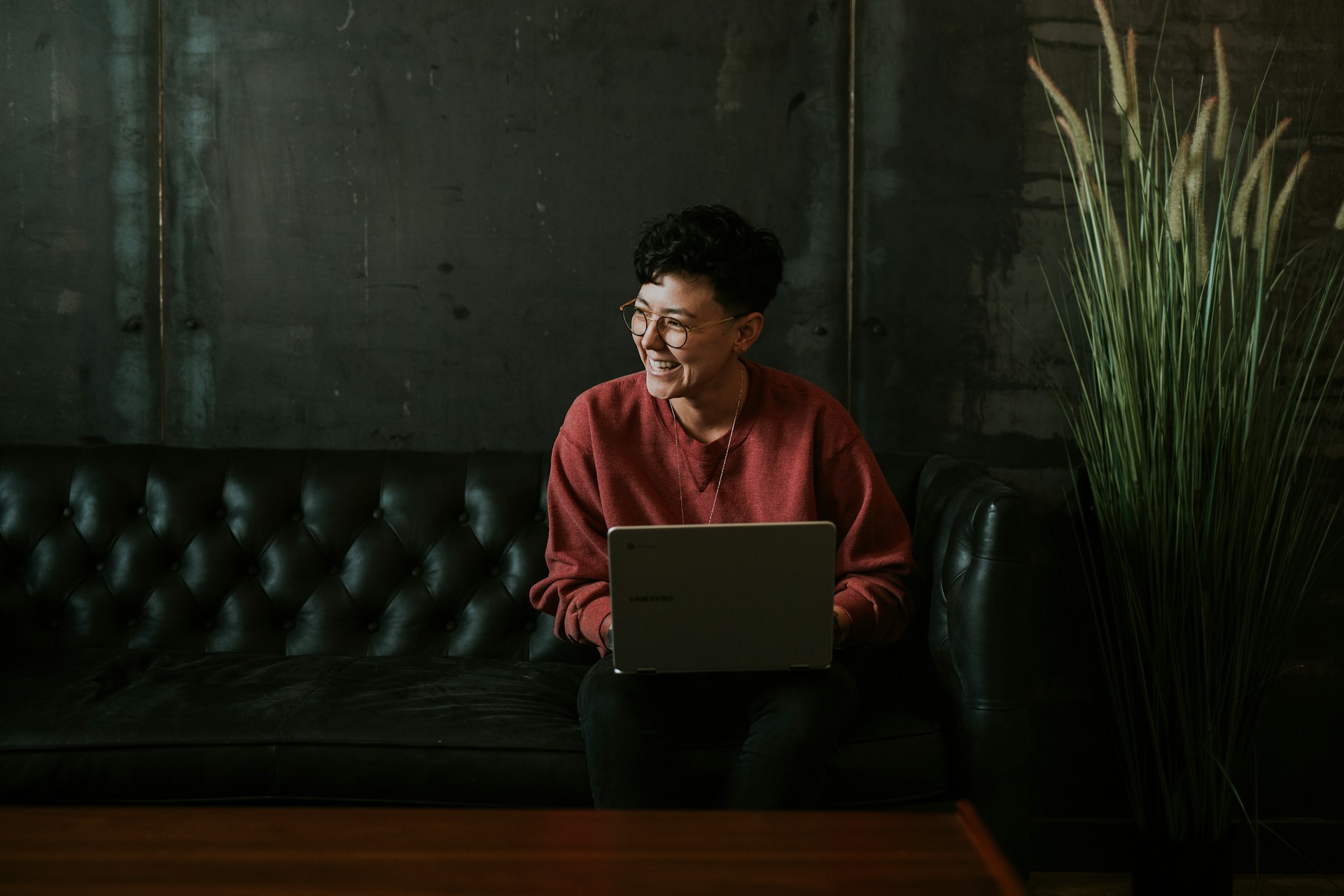 A person with short hair and glasses sits on a leather couch writing in a laptop and laughing at something off to the side.