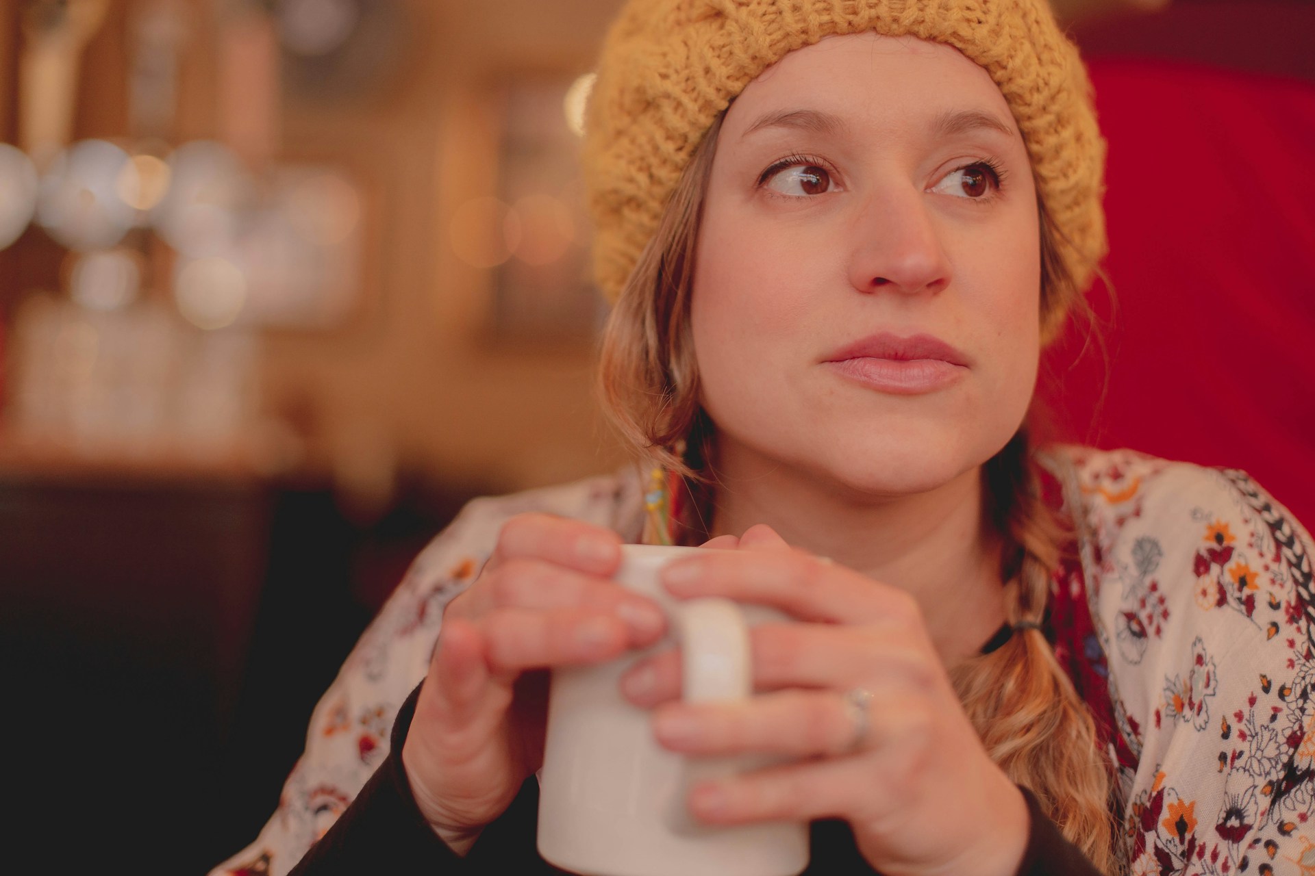 A person in a yellow beanie holds a mug in both hands while staring off into the distance.