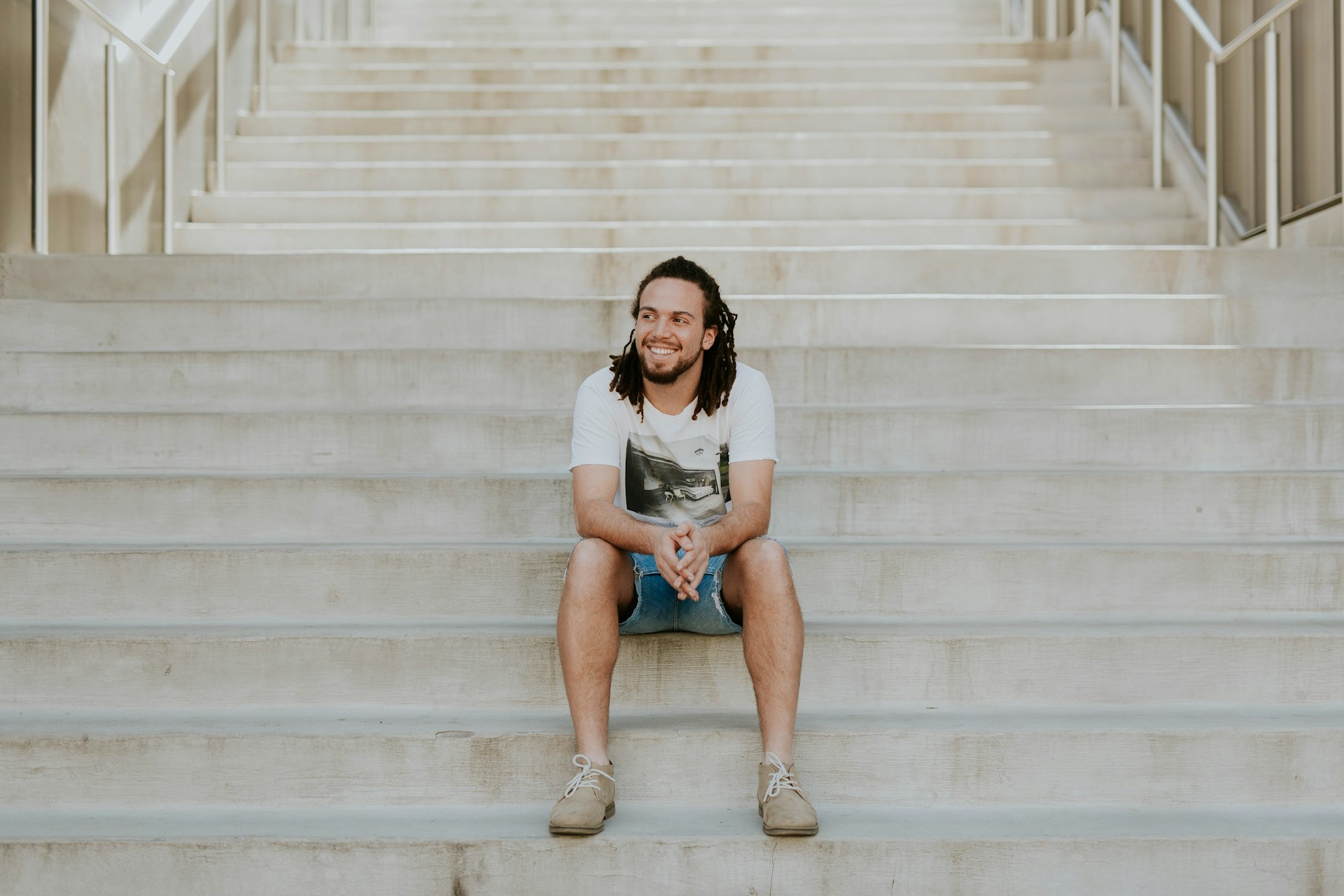 A person with dreadlocks casually sits on a staircase with their elbows resting on their knees and their hands lightly clasped.