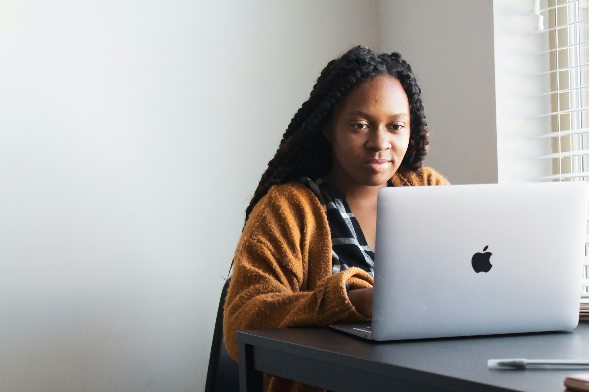 A person in an orange cardigan sits at a small table by a window, typing on a laptop.