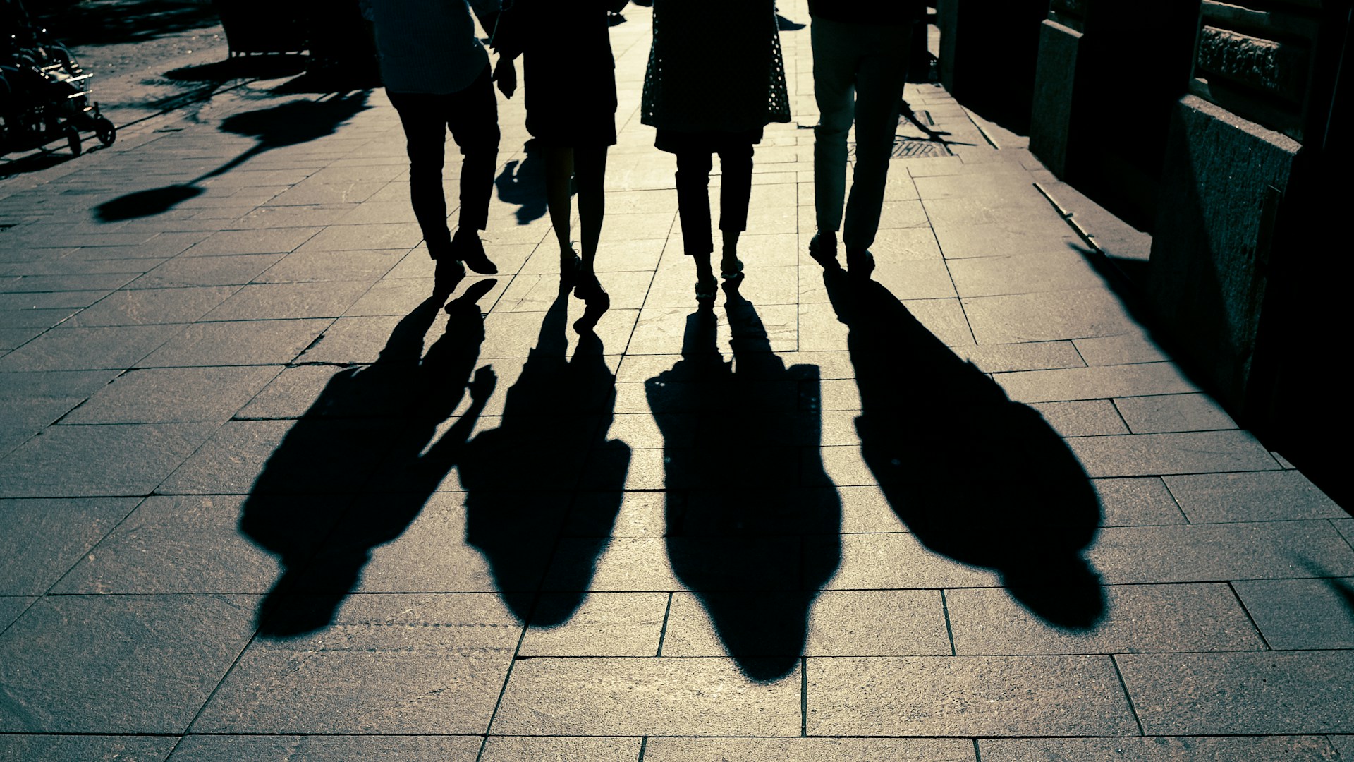 Four people walk along a wide sidewalk, casting long shadows on the ground.