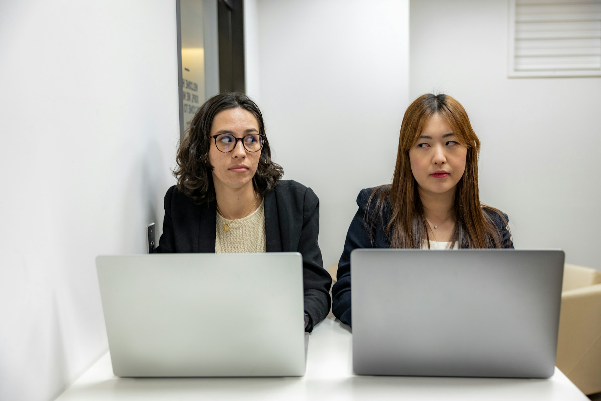 Two people sit side by side working on laptops and giving each other side-eye.e