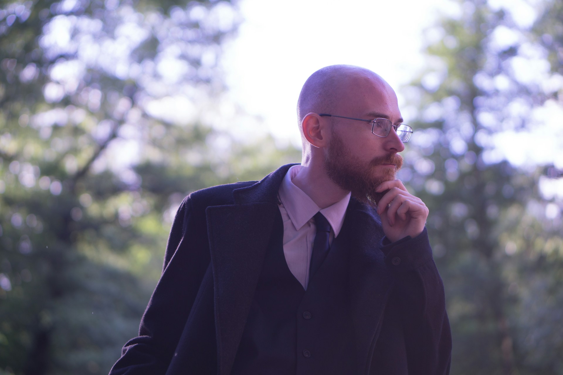 A bald person in a suit stands outside and touches their chin as they look off to the side, thinking.