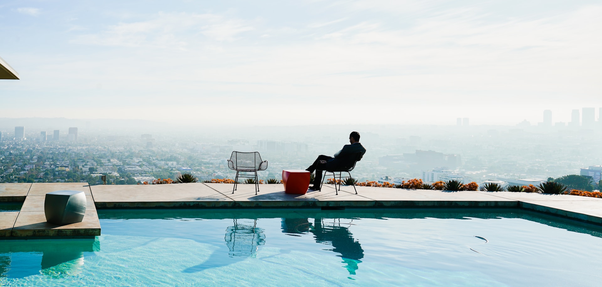 A wealthy person sits in a chair beside a pool at a fancy home, looking out over a city.